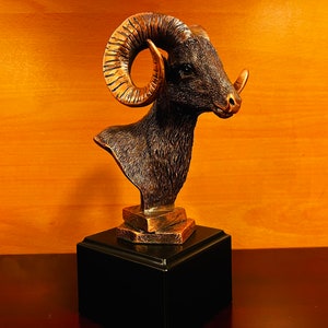 May include: A bronze bust of a ram with curled horns on a black base. The ram's head is turned to the right and its horns are curled forward. The bust is on a black base with a gold-colored platform.