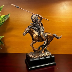 May include: Bronze-colored statue of a Native American warrior on horseback, holding a spear. The sculpture is mounted on a black rectangular base. The warrior is wearing a feathered headdress and traditional clothing. The horse is in mid-gallop.