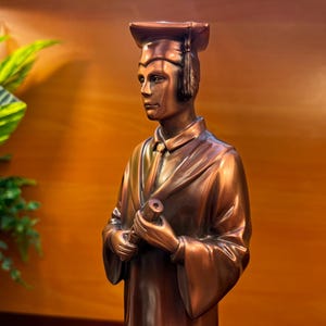 May include: Bronze-colored statue of a graduate in cap and gown, holding a rolled diploma. The figure has a serious expression and is set against a warm, wooden background. The statue is a decorative item.