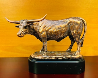 May include: A bronze sculpture of a longhorn steer with large horns, standing on a black base. The steer is depicted in a realistic pose with detailed features.