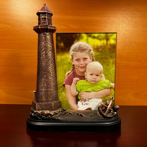 May include: A bronze lighthouse picture frame with a photo of two children, a girl and a boy, in a green field. The frame has a small anchor at the bottom and the text "5" x "7" Gwi" on the photo.