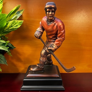 May include: A bronze sculpture of a hockey player in a red jersey, holding a hockey stick and wearing a helmet. The sculpture is on a black base with a bronze top.
