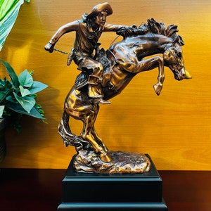 May include: A bronze sculpture of a cowboy riding a bucking horse. The cowboy is wearing a hat and holding onto the reins. The horse is rearing up on its hind legs. The sculpture is on a black base.