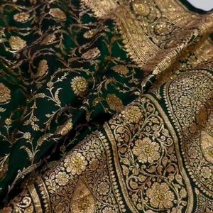 May include: A green and gold silk saree with intricate floral patterns and a border of gold thread. The saree is a traditional Indian garment worn by women.