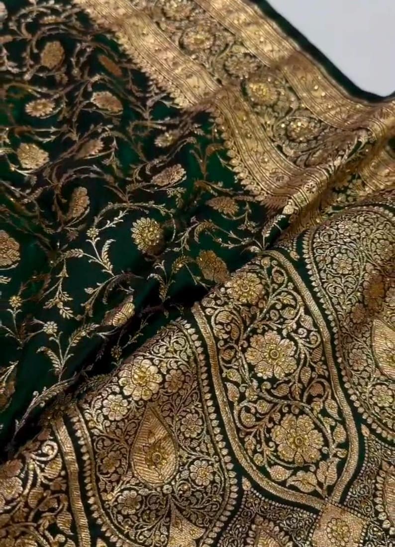 May include: A green and gold silk saree with intricate floral patterns and a border of gold thread. The saree is a traditional Indian garment worn by women.