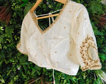 May include: Ivory-colored blouse with a V-neck and short sleeves. The blouse is embellished with gold-colored embroidery along the neckline and sleeve cuffs. Small gold-colored beads are scattered across the fabric. The blouse is hanging on a wooden hanger.
