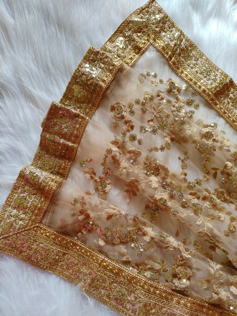 May include: A sheer gold and beige embroidered shawl with a gold sequin border. The shawl is made of a delicate fabric and has a floral design.