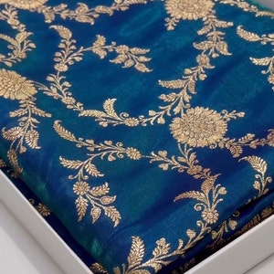 Könnte beinhalten: Ein blauer Seiden-Sari mit goldener Stickerei in einem Blumenmuster. Der Sari ist gefaltet und in einer weißen Schachtel verpackt.