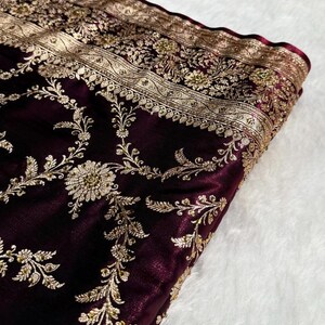 May include: Close-up of a deep burgundy fabric with intricate gold embroidery. The design features floral and vine patterns along the edges and throughout the material. The fabric has a soft, velvety texture.
