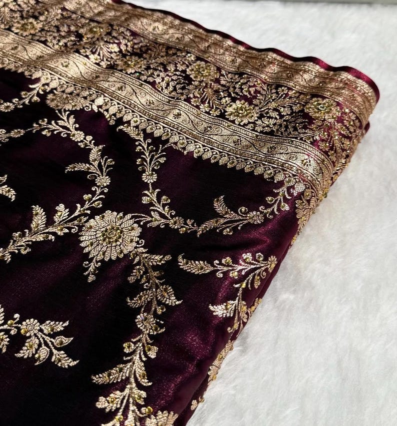 May include: Close-up of a deep burgundy fabric with intricate gold embroidery. The design features floral and vine patterns along the edges and throughout the material. The fabric has a soft, velvety texture.
