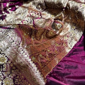 May include: Close-up of a luxurious, ornate textile in shades of deep purple, gold, and cream. The fabric features intricate floral and paisley patterns with shimmering embellishments, creating a rich and textured appearance. The design suggests a formal or ceremonial garment.