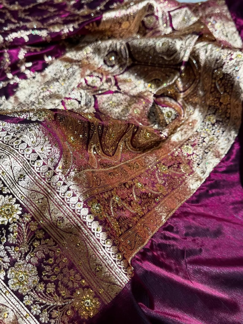 May include: Close-up of a luxurious, ornate textile in shades of deep purple, gold, and cream. The fabric features intricate floral and paisley patterns with shimmering embellishments, creating a rich and textured appearance. The design suggests a formal or ceremonial garment.