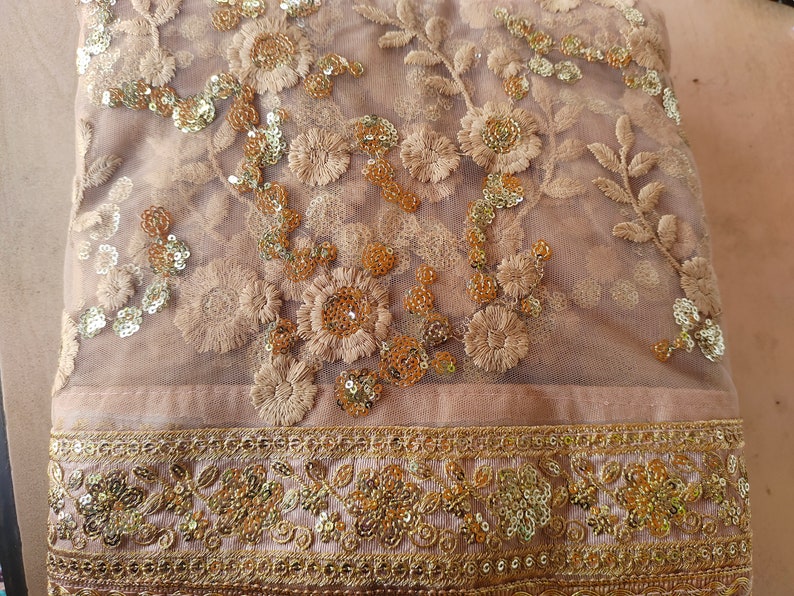 May include: A beige sheer fabric with gold embroidery and sequins. The fabric has a floral pattern and a gold border.