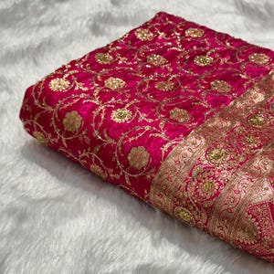 May include: A folded piece of vibrant fuchsia fabric with intricate gold embroidery. The design features floral patterns and ornate borders, creating a luxurious and detailed textile. The fabric is likely used for clothing or decorative purposes.
