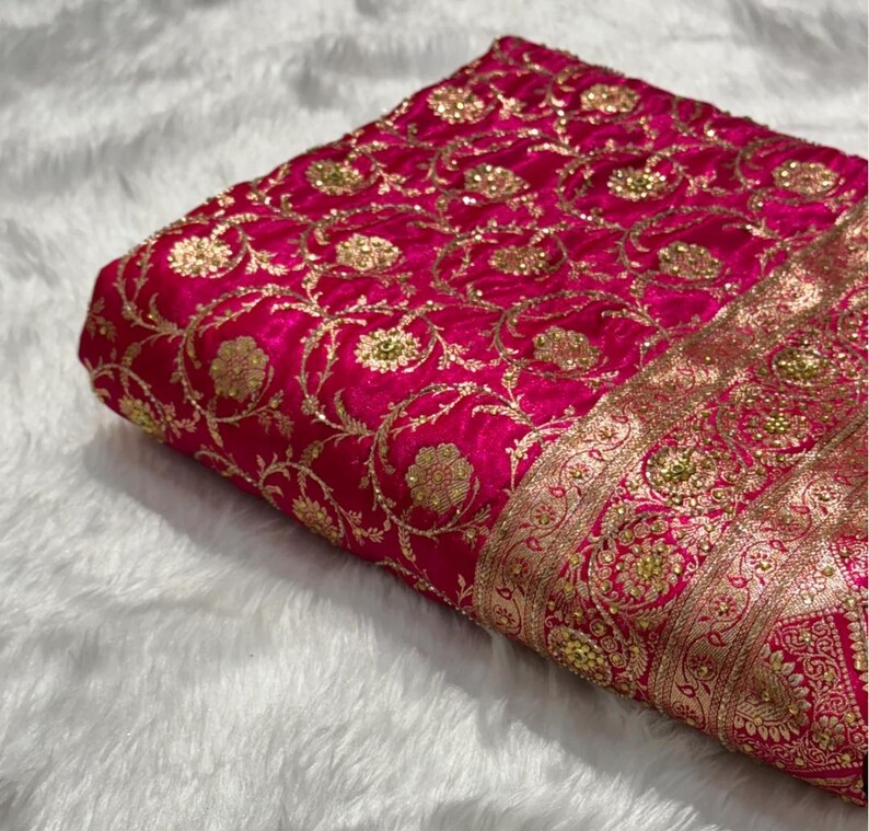 May include: A folded piece of vibrant fuchsia fabric with intricate gold embroidery. The design features floral patterns and ornate borders, creating a luxurious and detailed textile. The fabric is likely used for clothing or decorative purposes.