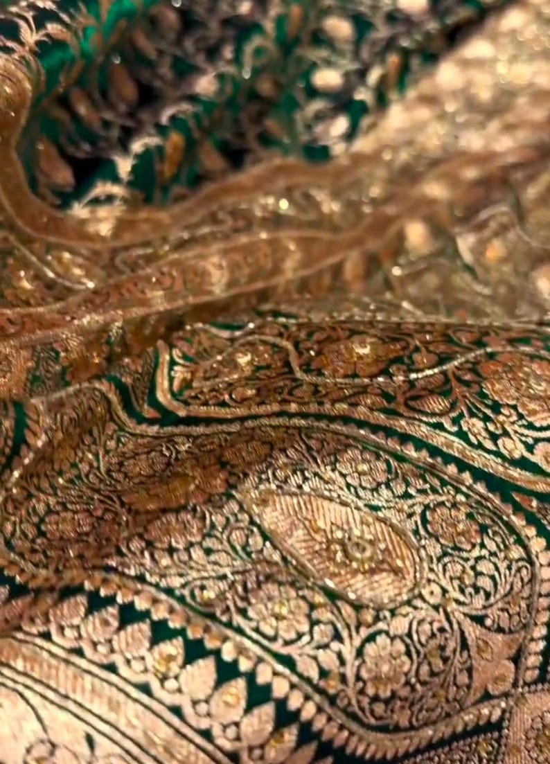May include: Close-up of a green and gold fabric with intricate paisley patterns. The fabric is richly embroidered with gold thread and sequins, creating a luxurious and ornate look.