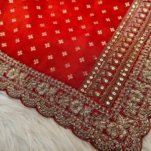 May include: A rich red textile with detailed gold embroidery and sequin embellishments. The fabric has a repeating pattern of small, gold floral designs. The edges are finished with a scalloped border, creating an elegant and ornate design.