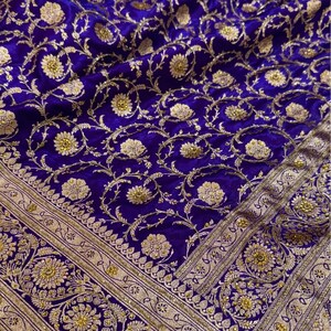 May include: Close-up of a luxurious, royal blue fabric with an intricate gold floral pattern. The design features detailed flowers and vines, with a decorative gold border. The fabric has a rich, velvety texture, suggesting a high-end textile.