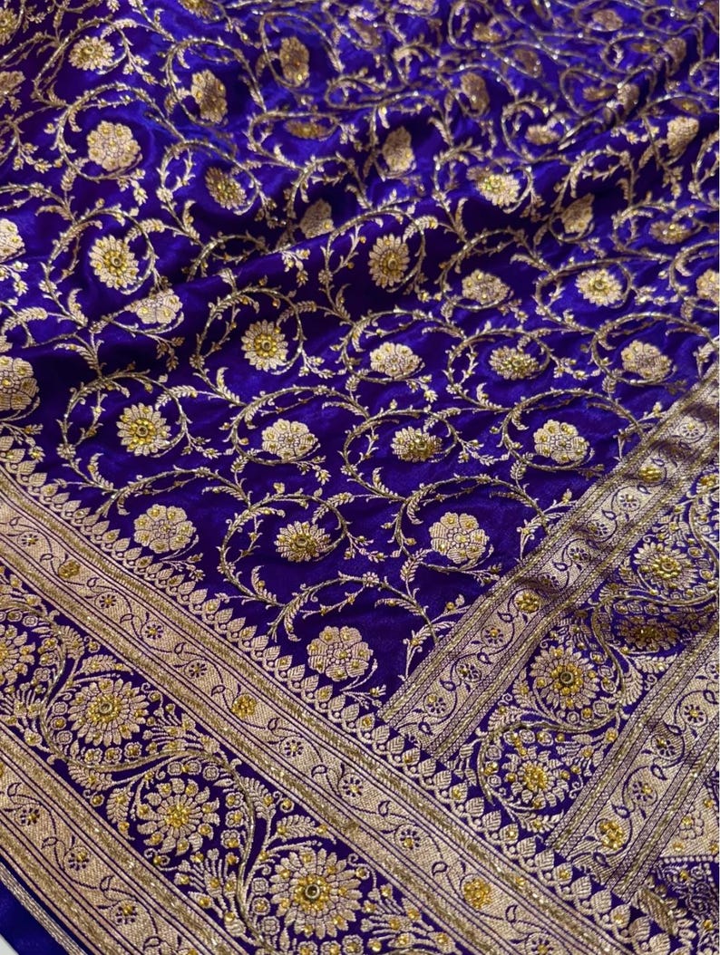 May include: Close-up of a luxurious, royal blue fabric with an intricate gold floral pattern. The design features detailed flowers and vines, with a decorative gold border. The fabric has a rich, velvety texture, suggesting a high-end textile.