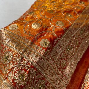 May include: Close-up of a vibrant orange textile with intricate gold embroidery. The fabric features floral and geometric patterns, with a detailed border. The design includes small, sparkling embellishments, creating a luxurious and ornate appearance.