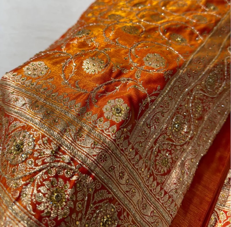 May include: Close-up of a vibrant orange textile with intricate gold embroidery. The fabric features floral and geometric patterns, with a detailed border. The design includes small, sparkling embellishments, creating a luxurious and ornate appearance.
