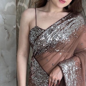 Könnte beinhalten: Eine Frau trägt ein braunes und silbernes, mit Pailletten besetztes Sari. Das Sari ist durchsichtig und hat ein zartes Muster. Die Frau trägt ein silbernes Armband am Handgelenk.