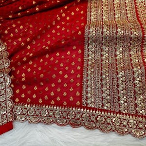 May include: A vibrant red fabric featuring a repeating pattern of small gold leaf motifs. The textile is bordered by a wide, intricate band of gold embroidery, sequins, and metallic thread, finished with a scalloped edge along the bottom.
