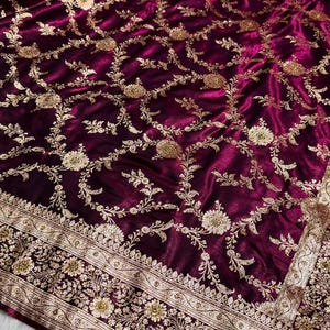 May include: A luxurious, deep burgundy fabric with intricate gold floral embroidery. The design features detailed vines and flowers, with a decorative border along the edge. The fabric has a rich, velvety texture.