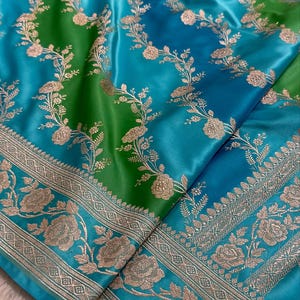 May include: A blue and green silk saree with a silver floral pattern. The saree has a border with a geometric design.
