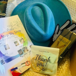 Può includere: Un cappello color turchese, un foglio di istruzioni 'Step By Step' e un piccolo sacchetto con la scritta 'Thank you!' sono visibili all'interno di una scatola. Sono inclusi anche un manuale per saldatore, un pennarello blu e una spilla dorata.