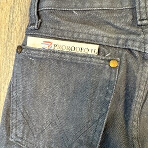 May include: Close-up of a pair of dark gray denim jeans with a back pocket. The pocket features a stitched design and a gold-colored button. A card with the text "PRORODEO HA" is tucked into the pocket.