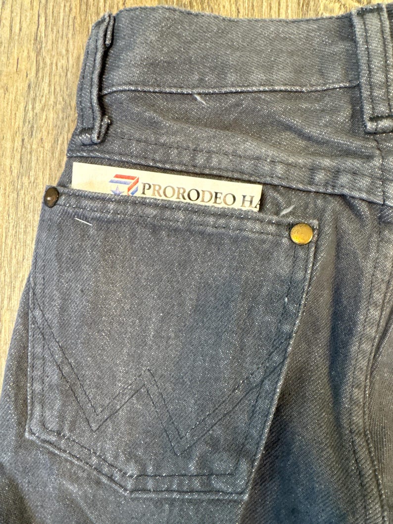 May include: Close-up of a pair of dark gray denim jeans with a back pocket. The pocket features a stitched design and a gold-colored button. A card with the text "PRORODEO HA" is tucked into the pocket.