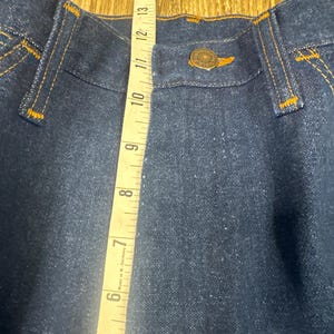K&ouml;nnte beinhalten: Nahaufnahme einer dunkelblauen Jeans mit orangefarbenen N&auml;hten. Ein gelbes Ma&szlig;band zeigt eine Messung von etwa 30 cm. Die Jeans hat einen Knopfverschluss und G&uuml;rtelschlaufen.