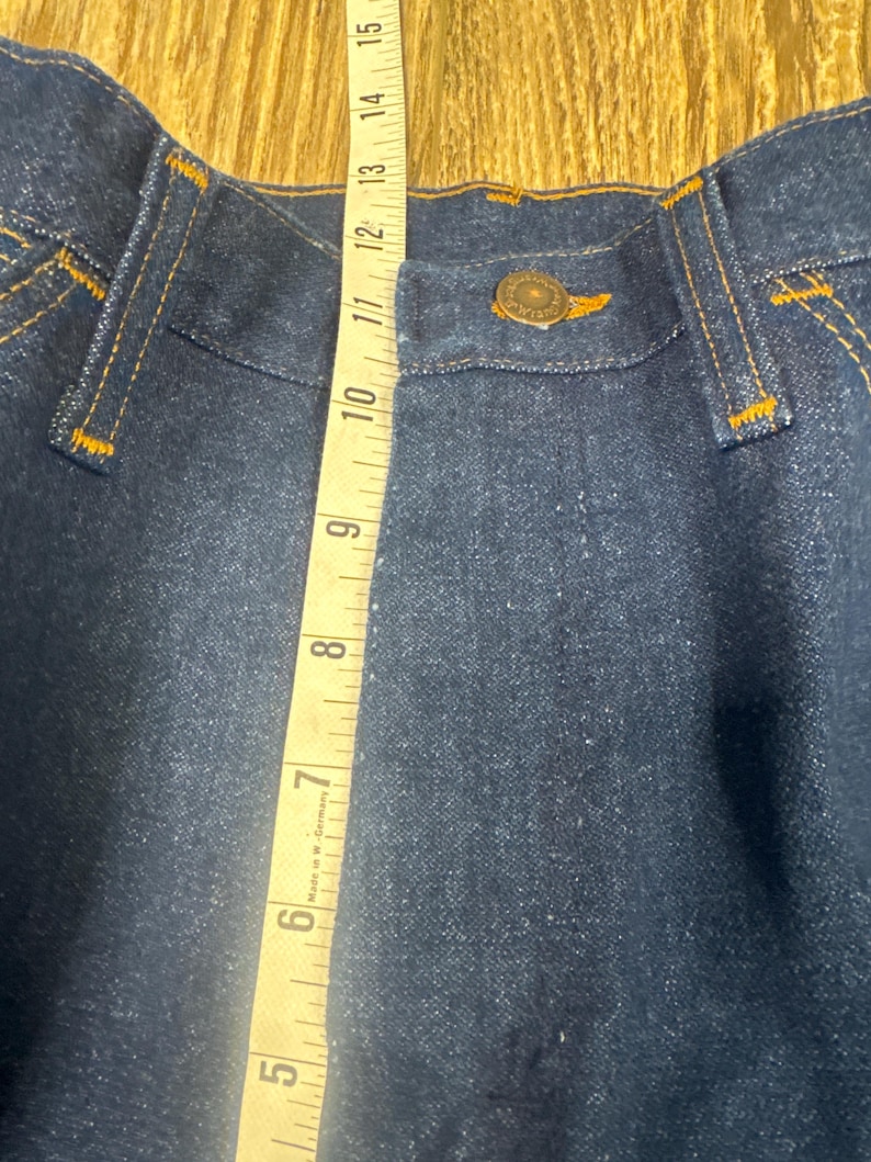 K&ouml;nnte beinhalten: Nahaufnahme einer dunkelblauen Jeans mit orangefarbenen N&auml;hten. Ein gelbes Ma&szlig;band zeigt eine Messung von etwa 30 cm. Die Jeans hat einen Knopfverschluss und G&uuml;rtelschlaufen.