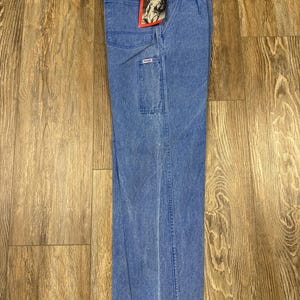 May include: Blue denim Wrangler work pants with multiple pockets and a straight leg. The pants have a faded wash and a Wrangler tag attached to the front. The pants are displayed on a wooden surface.