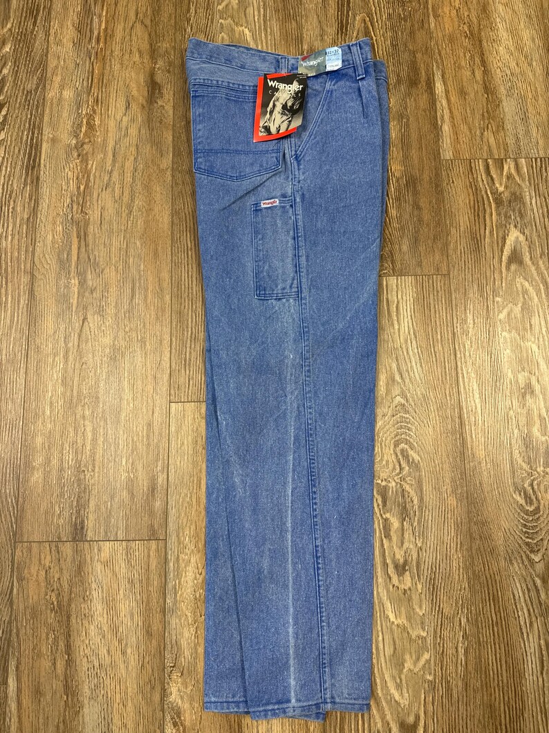 May include: Blue denim Wrangler work pants with multiple pockets and a straight leg. The pants have a faded wash and a Wrangler tag attached to the front. The pants are displayed on a wooden surface.