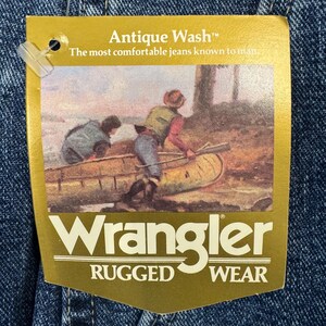 May include: Close-up of denim fabric with a gold tag. The tag says "Antique Wash" and "The most comfortable jeans known to man." The tag features a painting of two people in a canoe. The Wrangler logo is at the bottom, with "Rugged Wear" below.