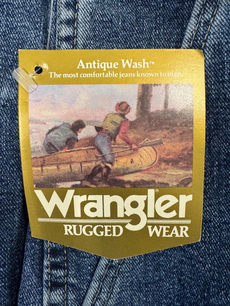 May include: Close-up of denim fabric with a gold tag. The tag says "Antique Wash" and "The most comfortable jeans known to man." The tag features a painting of two people in a canoe. The Wrangler logo is at the bottom, with "Rugged Wear" below.