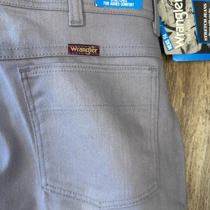 May include: Close-up of light gray Wrangler Flex-Fit waist jeans. The jeans have a back pocket and a brown Wrangler label. A blue and white tag reads "Flex-Fit Waist" and "Stretches for Added Comfort."