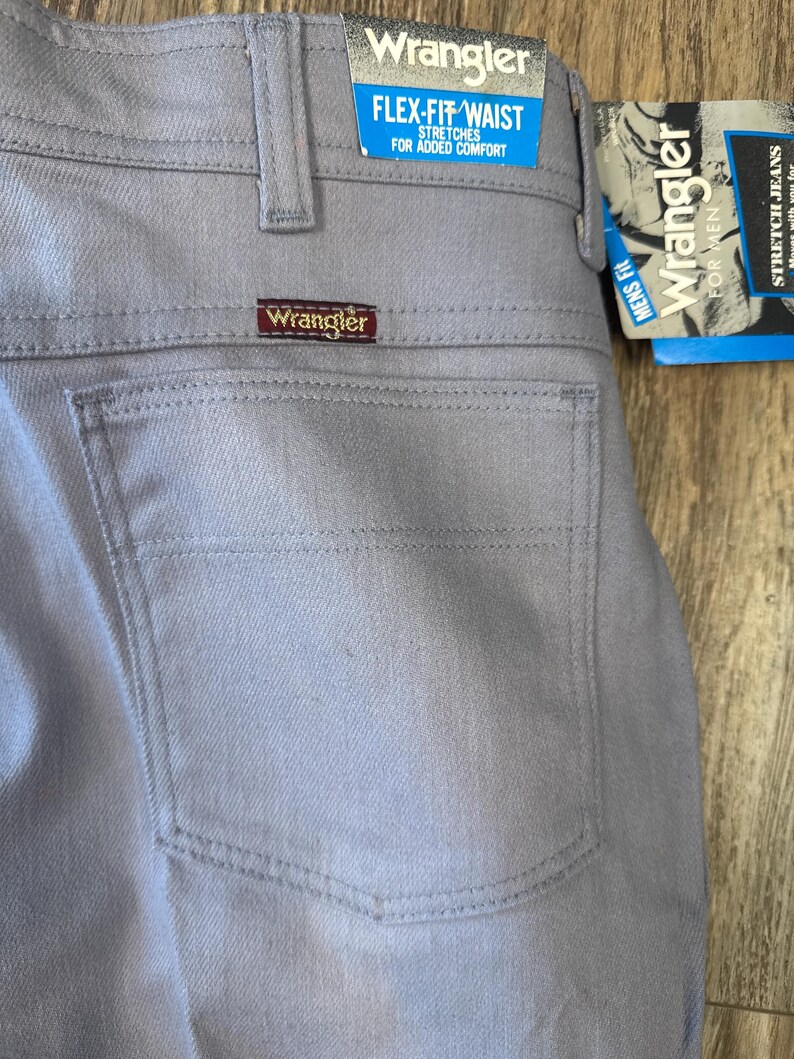 May include: Close-up of light gray Wrangler Flex-Fit waist jeans. The jeans have a back pocket and a brown Wrangler label. A blue and white tag reads "Flex-Fit Waist" and "Stretches for Added Comfort."