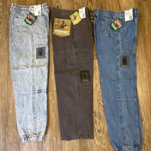 K&ouml;nnte beinhalten: Drei Wrangler Rugged Wear Cargo-Hosen in Acid-Wash-, Braun- und Blue-Denim. Jede Hose hat einen Aufn&auml;her und ein Etikett mit dem Wrangler-Logo. Die Hosen haben einen elastischen Bund und B&uuml;ndchen.