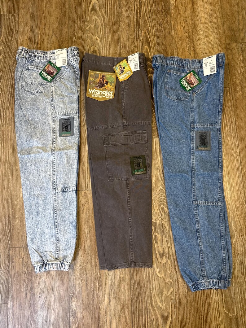 K&ouml;nnte beinhalten: Drei Wrangler Rugged Wear Cargo-Hosen in Acid-Wash-, Braun- und Blue-Denim. Jede Hose hat einen Aufn&auml;her und ein Etikett mit dem Wrangler-Logo. Die Hosen haben einen elastischen Bund und B&uuml;ndchen.
