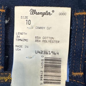 May include: Close-up of a pair of dark blue Wrangler denim jeans. The tag reads "Wrangler" and "SIZE 10, LENGTH 34". The tag also indicates the jeans are 65% cotton and 35% polyester. Made in the USA.