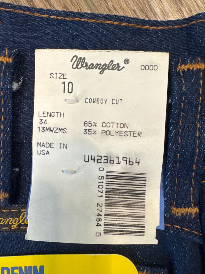 May include: Close-up of a pair of dark blue Wrangler denim jeans. The tag reads "Wrangler" and "SIZE 10, LENGTH 34". The tag also indicates the jeans are 65% cotton and 35% polyester. Made in the USA.