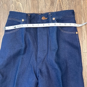 May include: Close-up of dark blue denim pants with a high waist. The pants have a button closure and pockets. A measuring tape shows the waist is approximately 14 inches across. The jeans have gold-colored rivets.