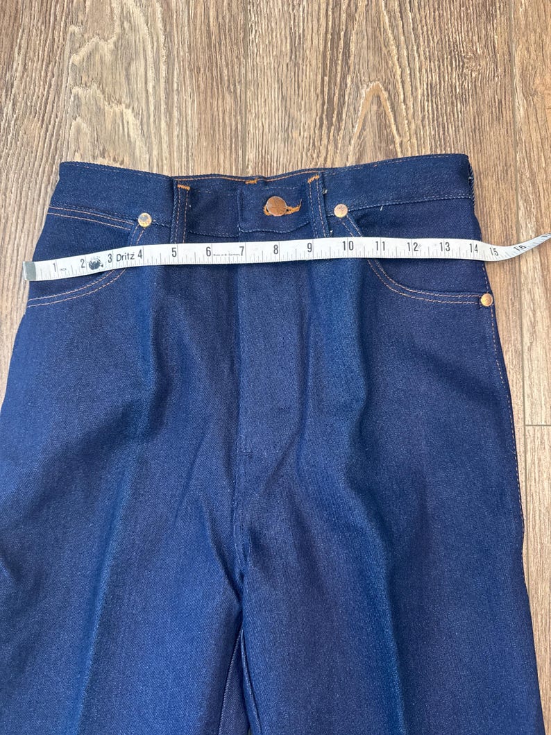 May include: Close-up of dark blue denim pants with a high waist. The pants have a button closure and pockets. A measuring tape shows the waist is approximately 14 inches across. The jeans have gold-colored rivets.