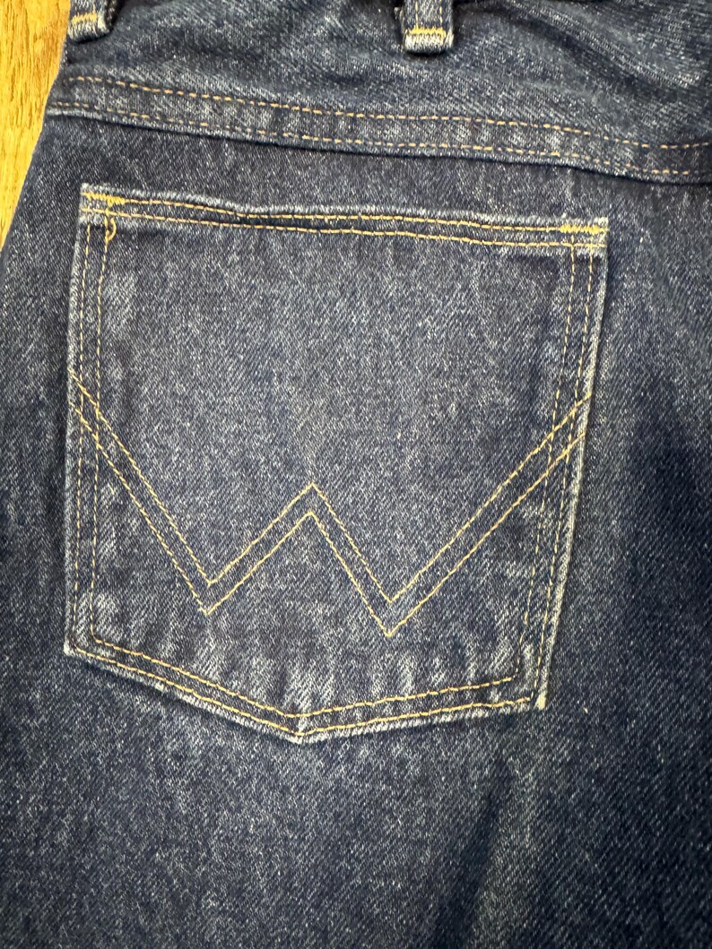 K&ouml;nnte beinhalten: Nahaufnahme einer dunkelblauen Jeans-Tasche mit gelber Naht. Die Tasche hat ein markantes gelbes "W"-Design. Der Denim hat eine strukturierte Optik.