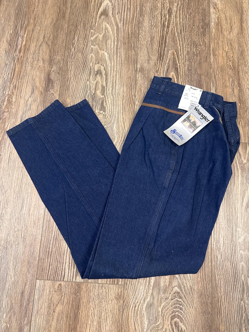 Puede incluir: Un par de pantalones vaqueros de mezclilla azul oscuro con un ribete de cuero marr&oacute;n en la cintura. Los pantalones tienen una etiqueta de Wrangler en el bolsillo trasero y una etiqueta de Wrangler m&aacute;s peque&ntilde;a en la parte delantera.
