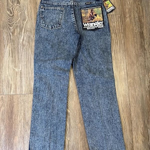Może przedstawiać: Niebieskie dżinsy Wrangler Rugged Wear. Dżinsy mają klasyczny pięciokieszeniowy design, proste nogawki i sprany kolor. Tylna kieszeń ma naszywkę Wrangler z grafiką osoby i napisem "Rugged Wear". Dołączona jest metka Wrangler.