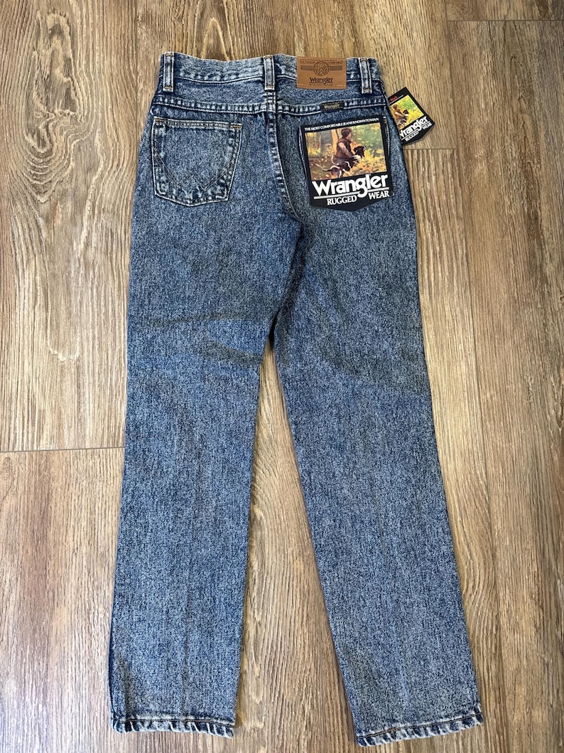 Może przedstawiać: Niebieskie dżinsy Wrangler Rugged Wear. Dżinsy mają klasyczny pięciokieszeniowy design, proste nogawki i sprany kolor. Tylna kieszeń ma naszywkę Wrangler z grafiką osoby i napisem "Rugged Wear". Dołączona jest metka Wrangler.